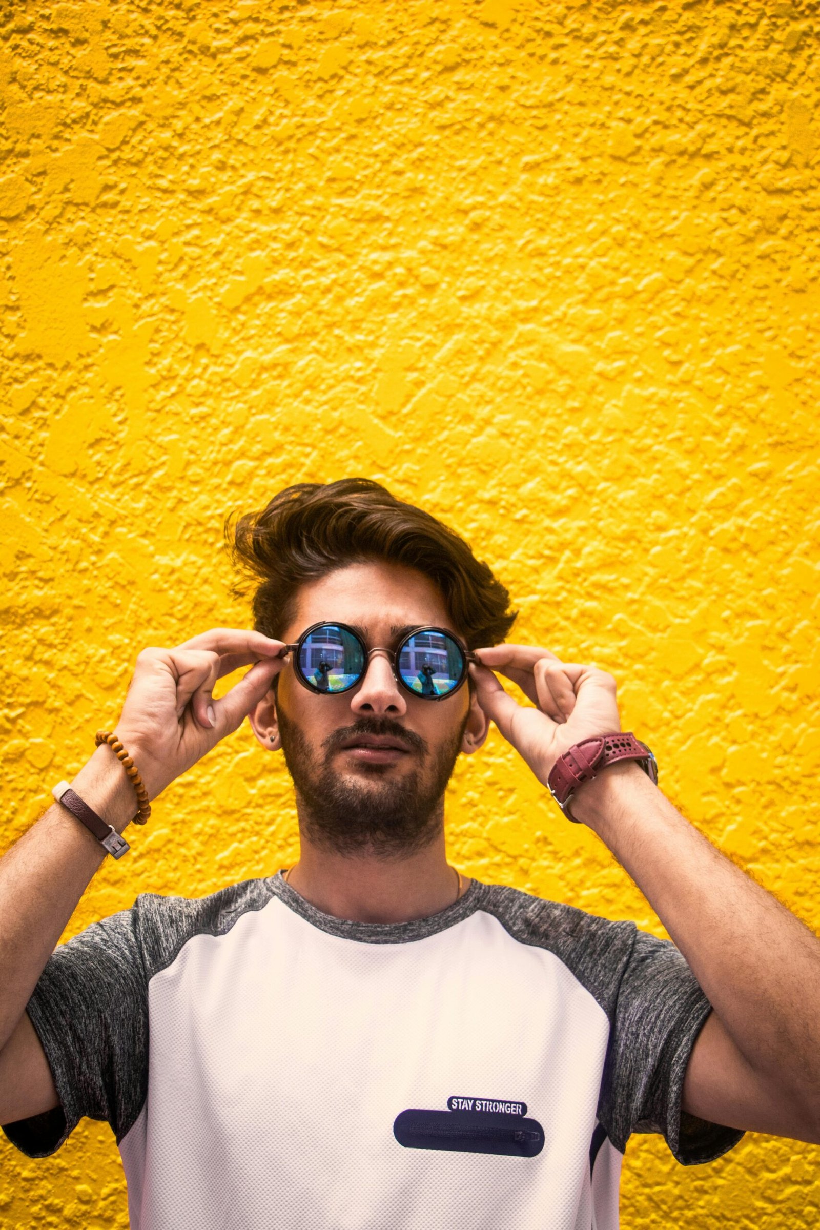 Young man with sunglasses standing against a vibrant yellow wall in daylight.