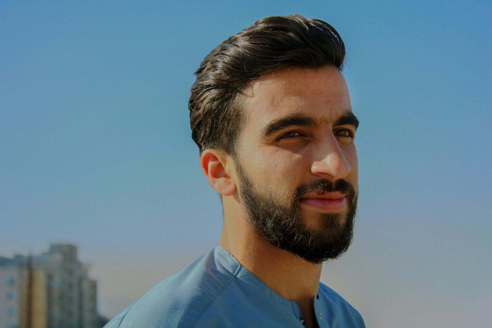 A close-up outdoor portrait of a young man with facial hair, exuding confidence under a clear sky.